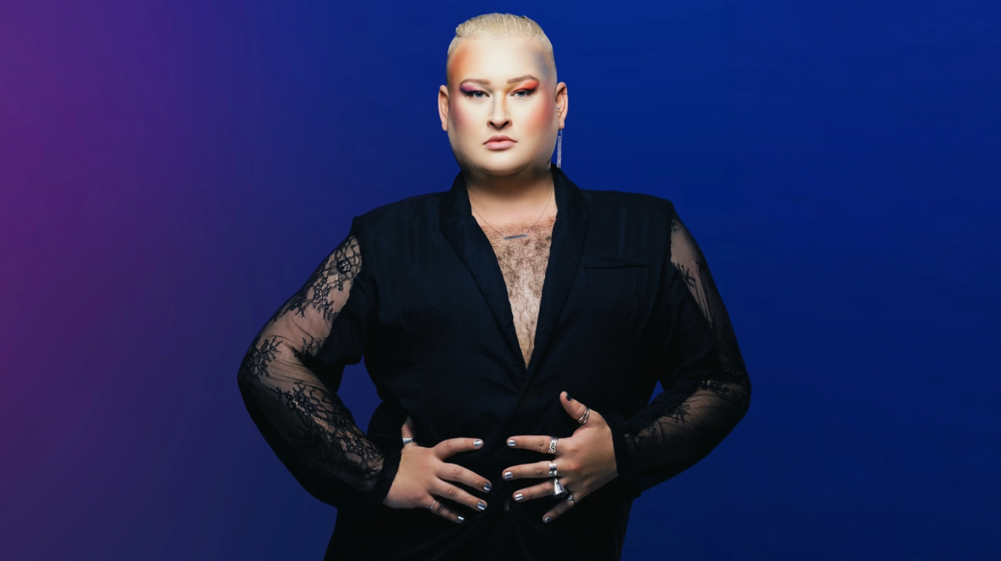 A person with short blonde hair and vibrant, multi-colored eyeshadow poses against a blue and purple gradient background. They are wearing a black blazer with lace sleeves and silver jewelry, including multiple rings, earrings, and a necklace, with their hands resting on their waist.
