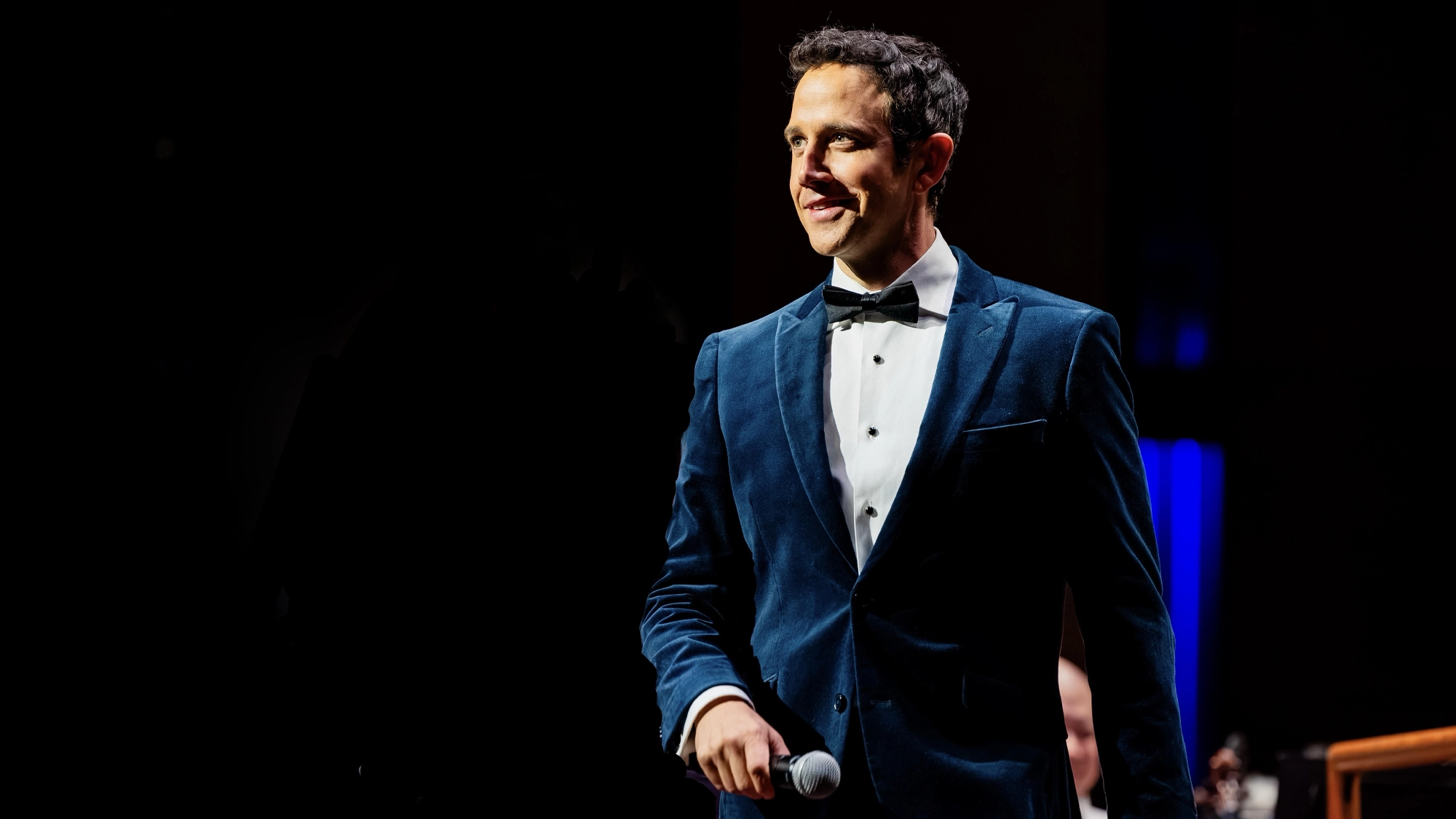 Santino Fontana, wearing a blue velvet tuxedo jacket and black bow tie, stands on a dark stage smiling toward the right while holding a microphone. He is spotlighted against a deep black background with faint blue lighting visible in the corner.