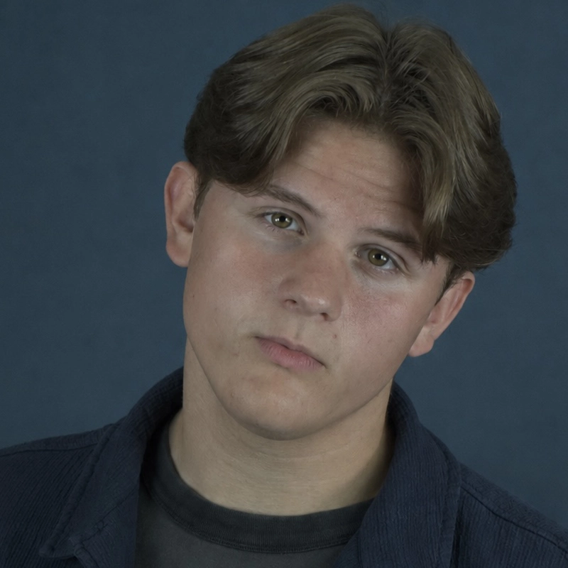 Josiah Schmidt looks directly at the camera with a serious expression. He has light brown hair parted in the middle and is wearing a dark grey t-shirt under a textured dark blue jacket against a solid blue background.