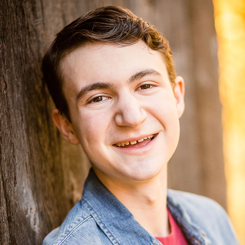 Tyler Cirulnick is a young man with brown hair who smiles warmly at the camera. He wears a blue denim jacket over a red shirt while leaning against a textured wooden surface.