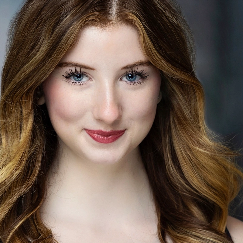 A headshot of Ryan Bagnolati-Camargo, a young woman with long, wavy brown hair with golden highlights. She has bright blue eyes, long eyelashes, and is wearing deep rose-colored lipstick. She is smiling gently against a mottled grey background.