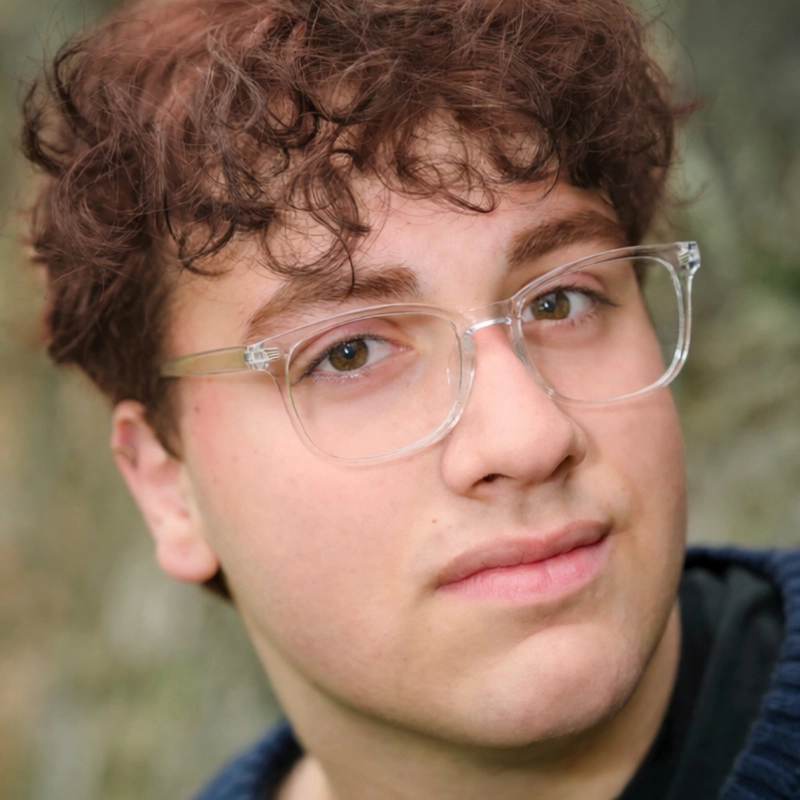 A young person with light skin, short curly brown hair, and clear eyeglasses looks into the camera with a slight smile. The background is blurred greenery; Dominick Stern wears a blue textured sweater over a black top.