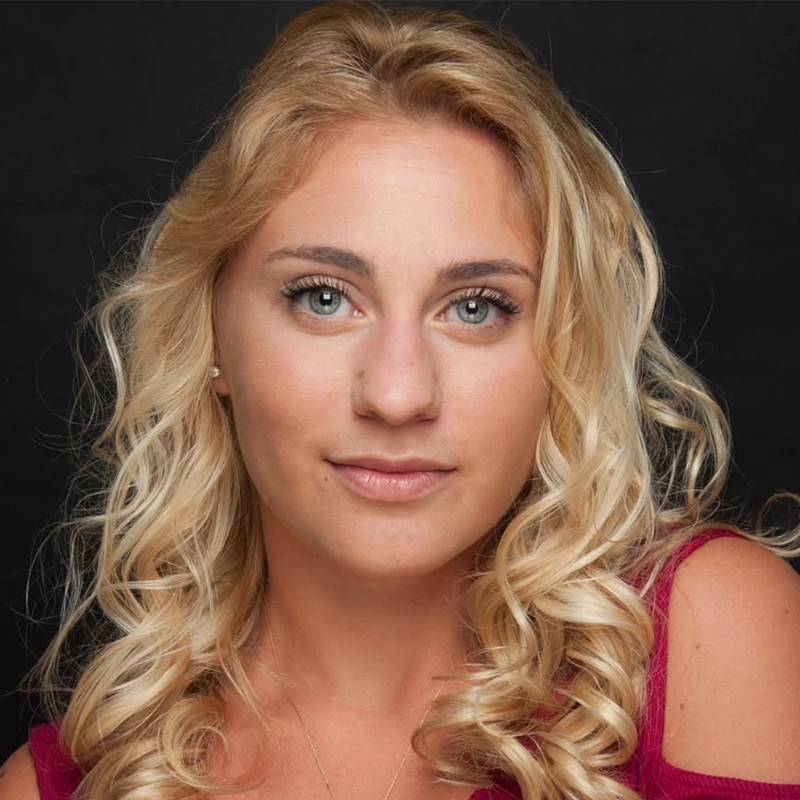 A headshot of Tyler Cirulnick, a woman with long, blonde curly hair and blue eyes looking directly at the camera. She is wearing a dark pink top against a black background.