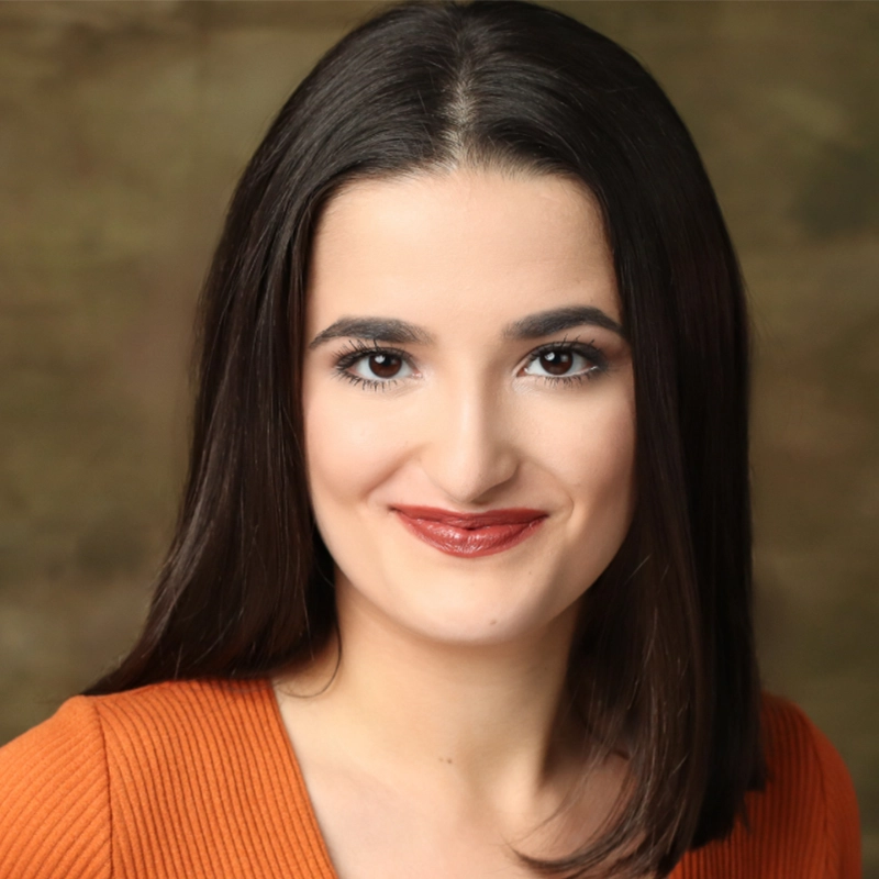 Tyler Cirulnick smiles at the camera with long dark hair. She wears a rust-colored top against a mottled, earth-toned background.