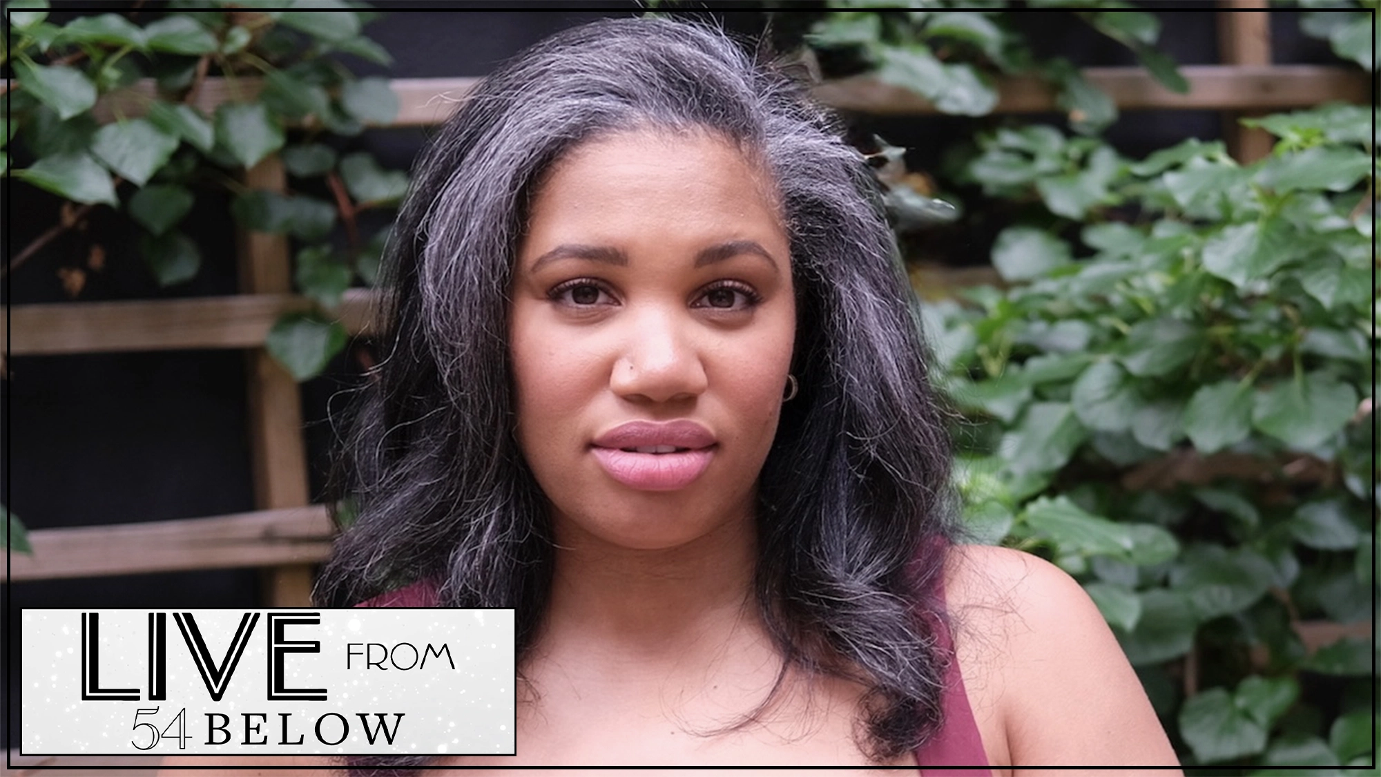 A woman with long, wavy, gray-streaked hair and a maroon top stands outdoors in front of green foliage, gazing at the camera. In the lower-left corner, a “LIVE FROM 54 BELOW” LIVESTREAM logo appears. Tara Jackson presents “Introduce Myself.”.