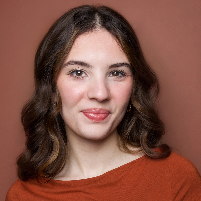A young woman with wavy brown hair smiles gently at the camera. She wears a burnt orange top and gold earrings. Her skin is fair, and she has light makeup. The background is a solid, warm brown color, complementing her shirt and overall tones.