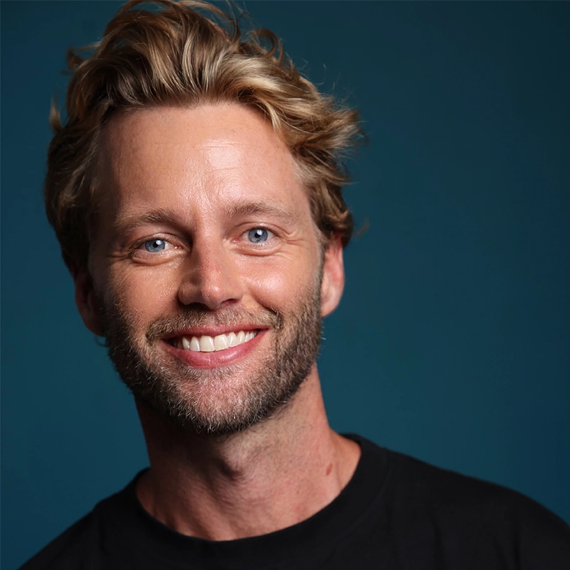 A close-up portrait of the smiling choreographer Trent Whiddon, featuring messy blonde hair, bright blue eyes, and light stubble. He wears a plain black crew-neck shirt against a solid, dark teal background.