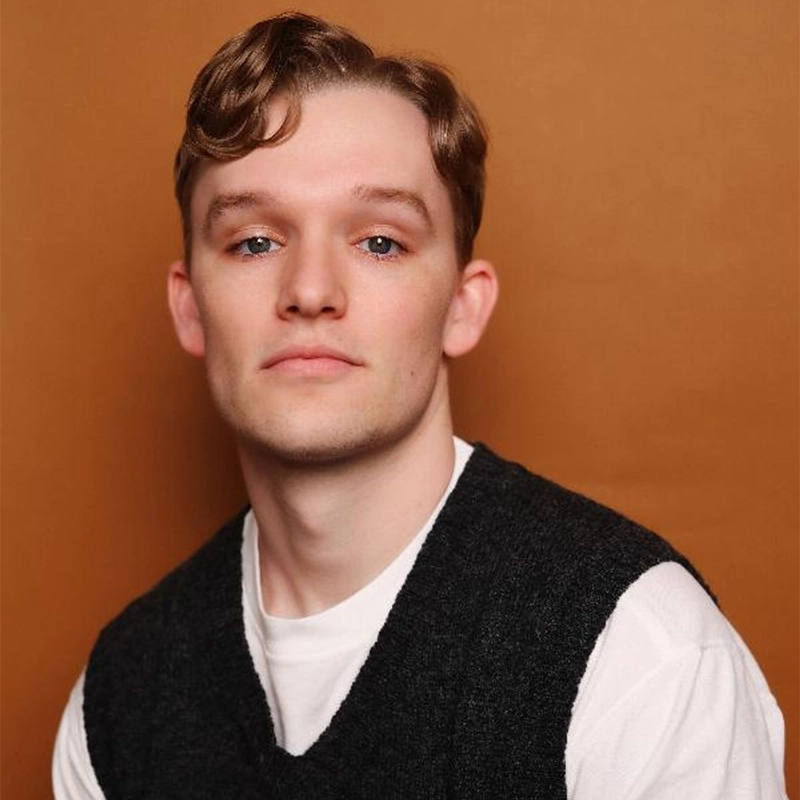 A head-and-shoulders portrait of Colin Trudell, a fair-skinned person with short, wavy reddish-brown hair, looking directly at the camera with a neutral expression. They wear a dark grey sweater vest over a white t-shirt against a solid, warm terracotta-colored background.