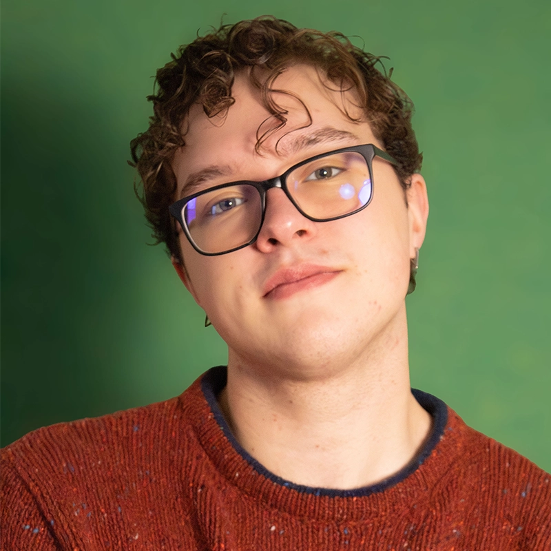 Tyler LaPointe, a young person with curly brown hair and glasses, looks toward the camera against a green background. They are wearing a textured, rust-colored knit sweater with a visible dark blue collar.