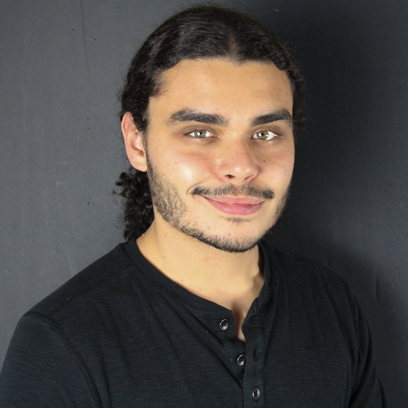 Keiran Varian is a young man with light skin, hazel eyes, and a light beard posing against a charcoal gray background. He has long, dark curly hair pulled back, wearing a black henley shirt while looking directly at the camera with a subtle, friendly smile.