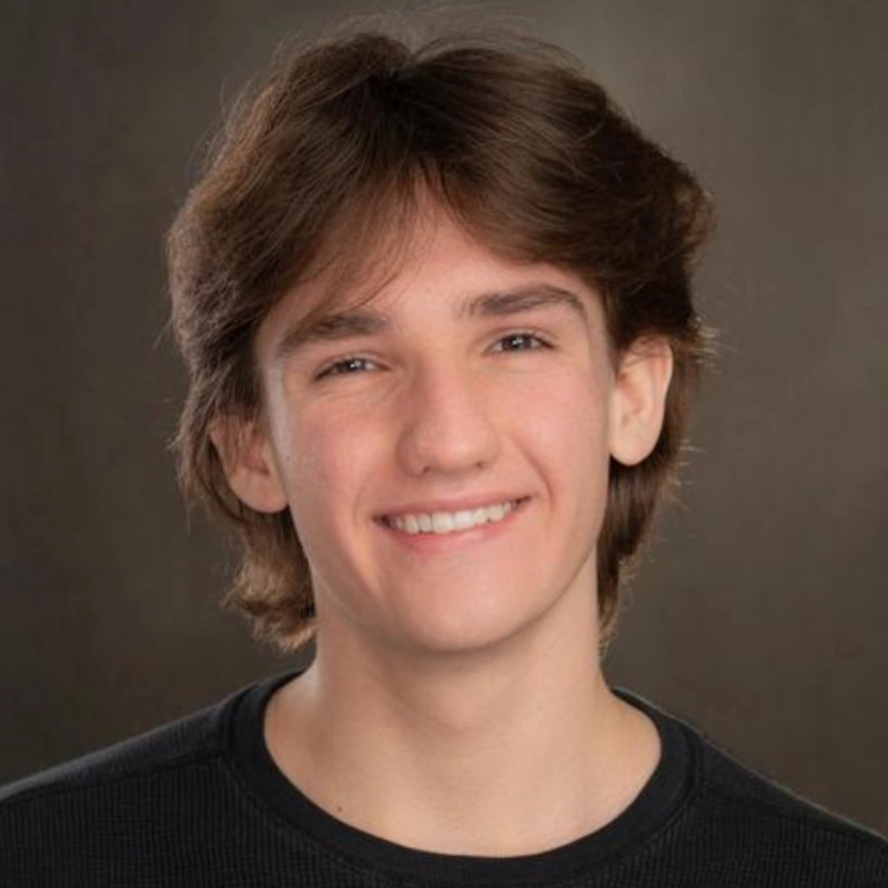 A portrait of Ryan Bagnolati-Camargo, a young person with chin-length, wavy brown hair and a warm smile. They are wearing a black crew-neck shirt against a neutral, mottled brown background.