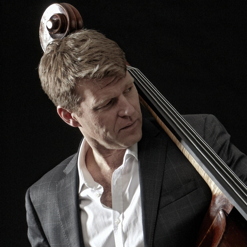 A fair-skinned man with short blonde hair looks intently to his right while holding an upright bass. He is wearing a dark, subtle-checkered suit jacket over an unbuttoned white dress shirt against a plain black background.