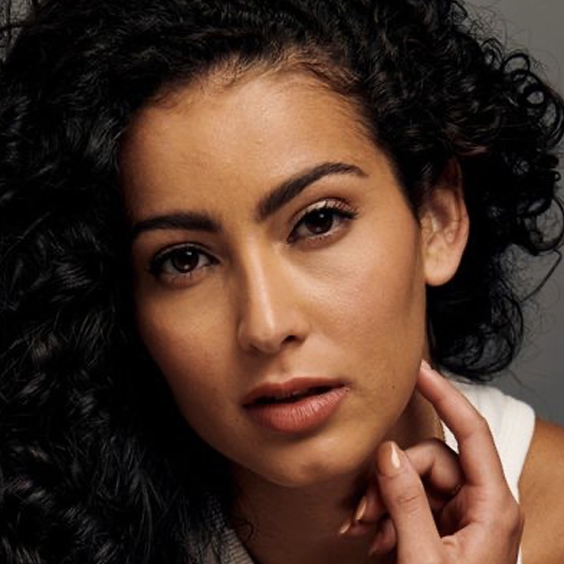 A portrait of Valeria Yamin with curly dark hair looking directly at the camera. She has a neutral expression, light brown skin, and is wearing a white top, with one hand gently touching her chin.