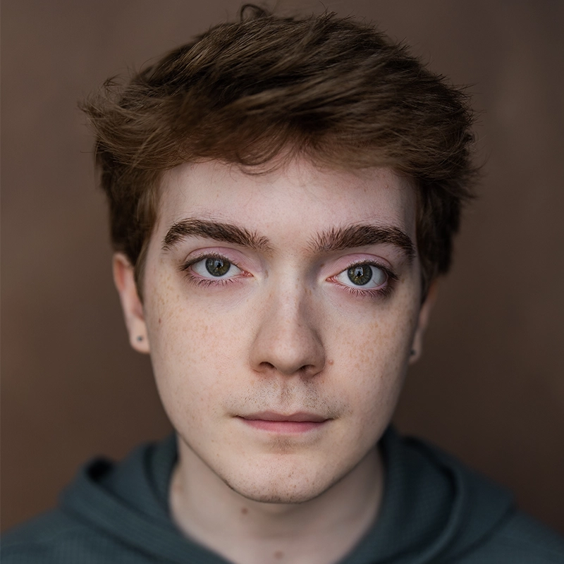 Actor Zell Steele Morrow in a headshot, wearing a dark hooded sweatshirt. They have short brown hair, light freckles, and a neutral expression while looking directly at the camera.