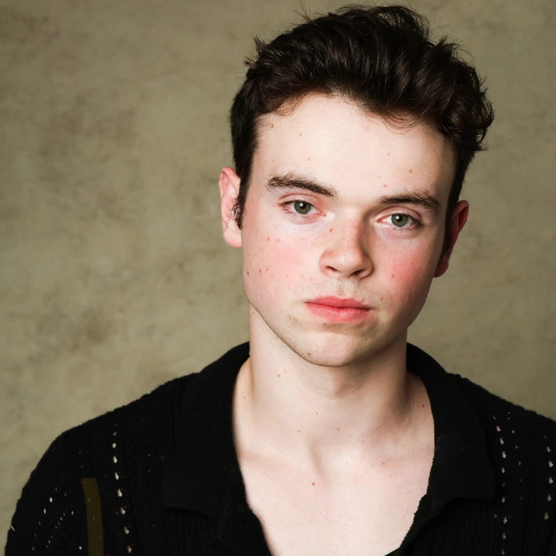 Eli Zimmerman, a young man with pale skin, dark wavy hair, and green eyes, stares directly at the camera. He wears a black textured sweater against a neutral backdrop, his expression neutral with some redness and faint freckles visible.