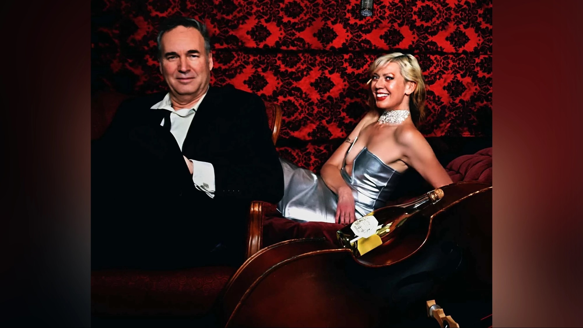 A man in a black suit and a woman in a silver dress lounge on a velvet couch against a red damask wallpaper background. A bottle of Champagne Bruts rests on the body of a double bass positioned in the foreground.