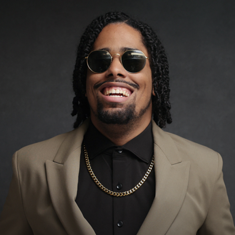 A smiling man with locs wears round sunglasses, a black shirt, a gold chain, and a tan blazer.